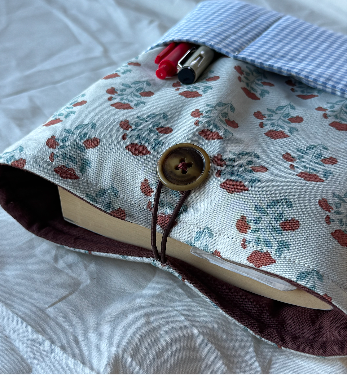 A fabric book cover or notebook sleeve with a floral print, shown holding a book on a wrinkled white sheet. The cover has a blue-and-white gingham pocket with pens in it, a brown button closure, and a dark brown elastic strap.