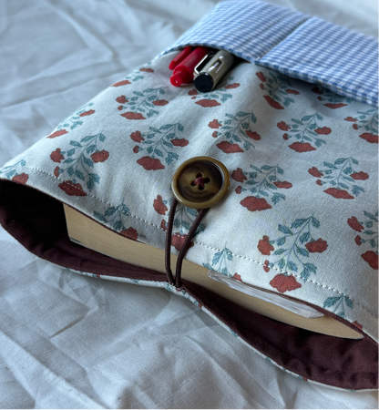 A fabric book cover or notebook sleeve with a floral print, shown holding a book on a wrinkled white sheet. The cover has a blue-and-white gingham pocket with pens in it, a brown button closure, and a dark brown elastic strap.